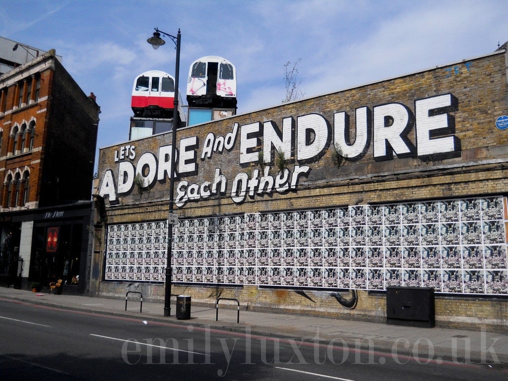 long brick wall with with two underground trains on top and large painted white words reading: "Let's Adore and Endure Each Other" - East London street art walk 