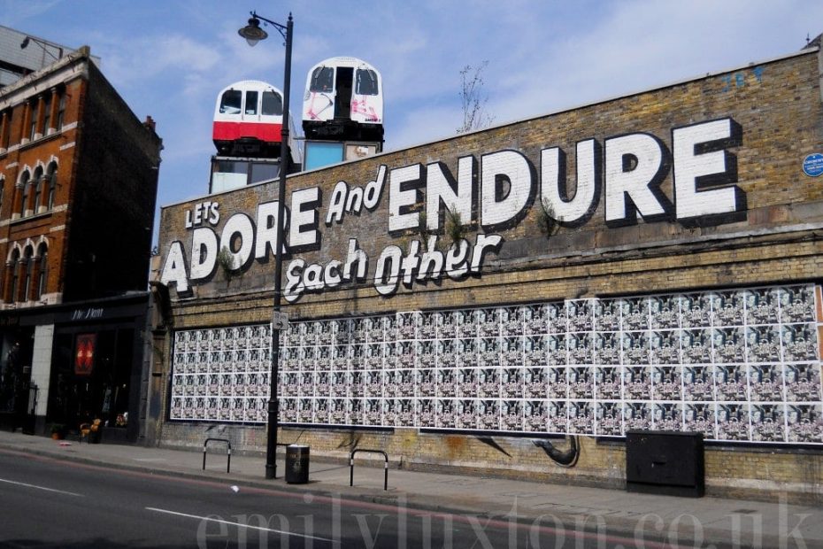 long brick wall with with two underground trains on top and large painted white words reading: "Let's Adore and Endure Each Other" - East London street art walk