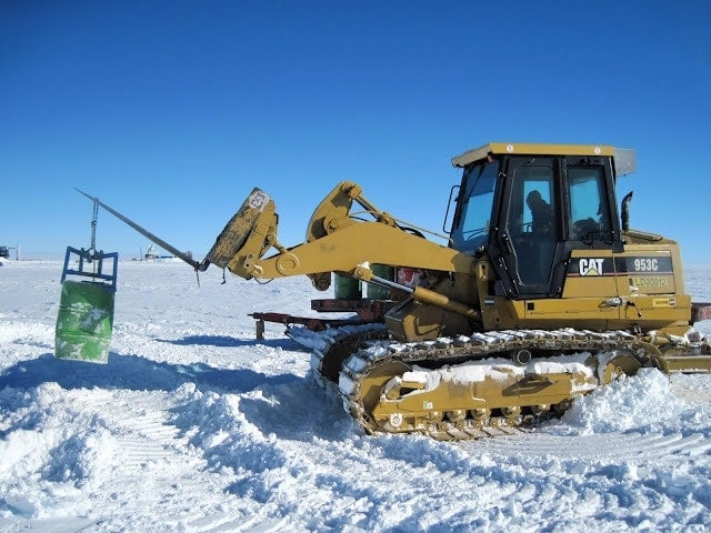 yellow CAT tractor driving on a large flat snowfield carrying a green metal drum under a bright blue sky - working in antarctica