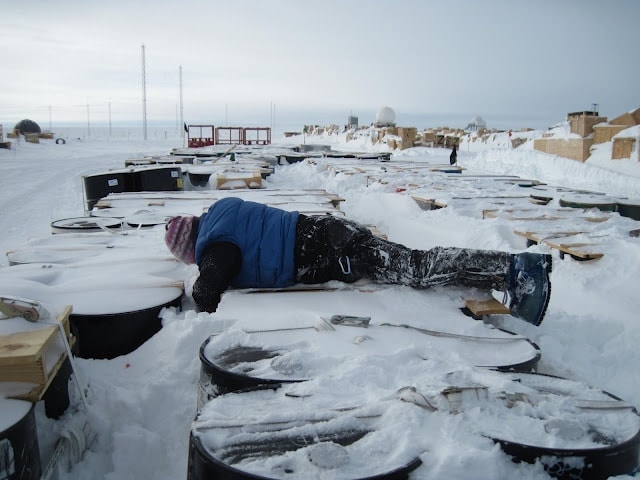 a woman wearing black trousers and a blue puffer jacket with a multicoloured wooly hat is lying face down on top of some large metal barrel drums covered in ice with snow-covered machinery in the background 