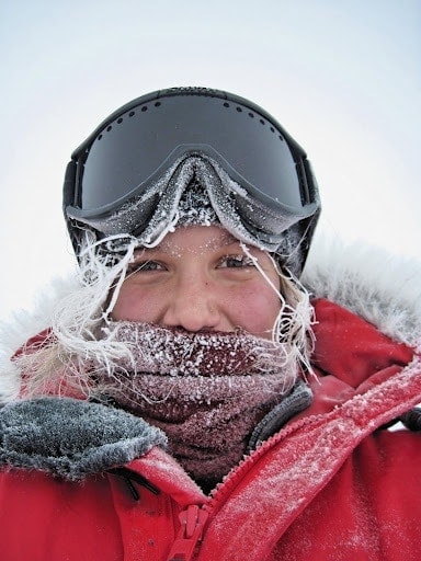 Portrait of Kiell, a girl who spent two summers working in Antarctica. Her fair hair is completely covered with white ice, she is wearing a brown scarf pulled up to her nose, with large black goggles pushed up on her head and a bright red coat with a fur lined hood.
