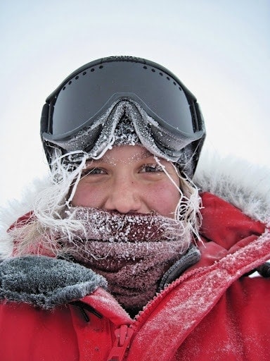 Portrait of Kiell, a girl who spent two summers working in Antarctica. Her fair hair is completely covered with white ice, she is wearing a brown scarf pulled up to her nose, with large black goggles pushed up on her head and a bright red coat with a fur lined hood. 