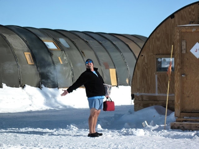 a man wearing blue shorts and a black fleece jumber with a goatee beard and a cigarette in his mouth holding a small red holdall in one hand and waving with his other, standing on a snowfiled in front of a  grey, metal, cylindrical shaped military-style building