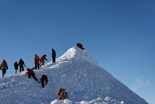 a line of small human figures silhouette against the white ice and blue sky climbing a conical shaped hill of snow and ice in antarctica