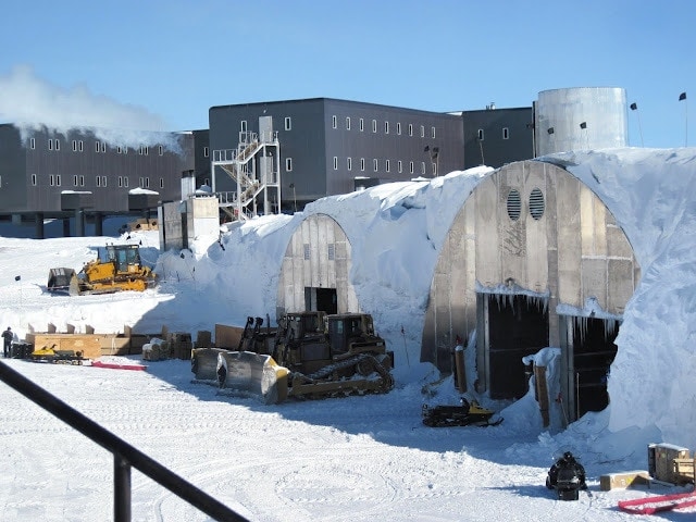 cylindrical hangers with clear fronts covered in snow with grey rectangular buildings in the background at the south pole station with snow in front and a clear blue sky overhead