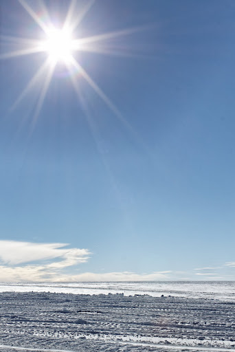 flat grey and white icy and snowy ground wiht nothing on it beneath a lot of empty blue sky with the high sun creating a star-shaped lens flare