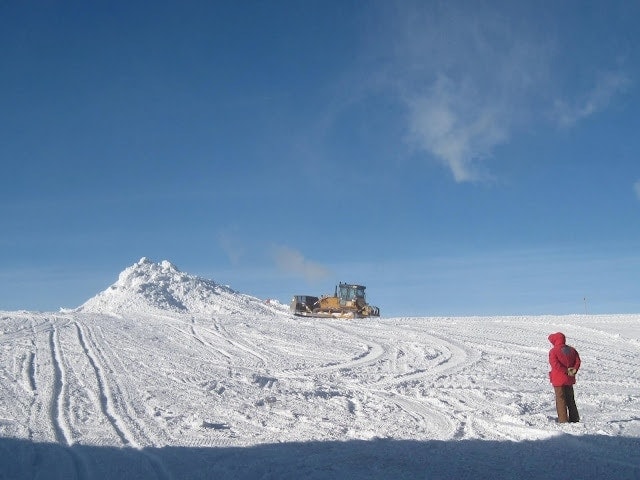 Watching a tractor push snow human figure facing away from the camera wearing brown trousers and a red hooded jacket standing on a large snowscape with a small snowy hill and a yellow tractor in the distance under a bright blue clear sky