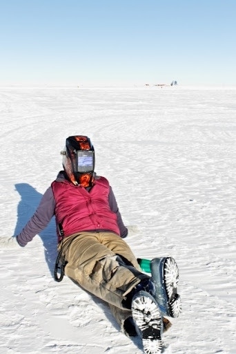 Kiell wearing light brown walking trousers, snow boots, and a red gilet over a long sleeved black top, with a full-face glass mask over her face, sitting on a snowfield to watch the solar eclipse
