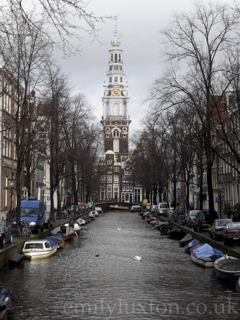 View along a canal towards the red brick, white topped tower of the Zuiderkerk church on a grey day with bare trees either side of the canal