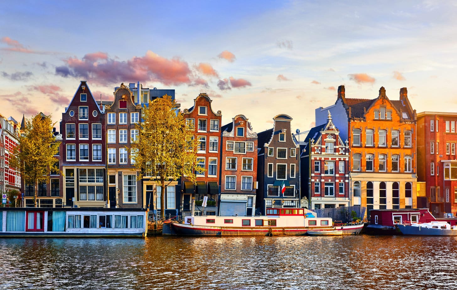 View of terraced townhouses across a river in Amsterdam Netherlands around sunset in spring with a canal boat on the watyer in front. One day in Amsterdam. 