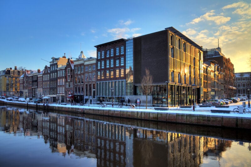 view of a square stone builing with a modern glass extension next to a canal with both buildings reflected in the water - the anne frank house and museum in amsterdam
