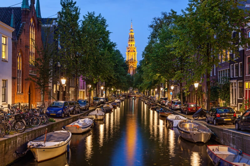 View along a canal from a bridge at night with boats moored along either side of the canal and a lit up tower at the end of the canal