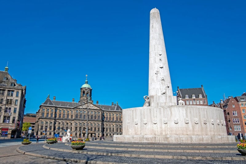 large white stone monument with a tall stone cilindar rising up towards a clear blue sky in a cobbled stone public square with a large stone palace out of focus behind