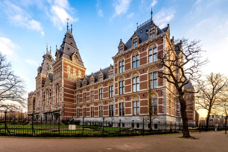 Exterior of a grand old palace built from red brick with beige trim on a sunny day in amsterdam with blue sky above