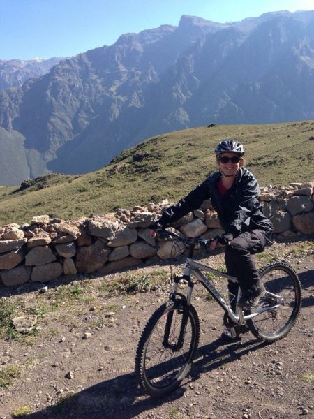 emily wearing a black raincoat on a bike cycling down a dirt path next to a stone wall with mountains  behind her and blue sky overhead on a sunny day