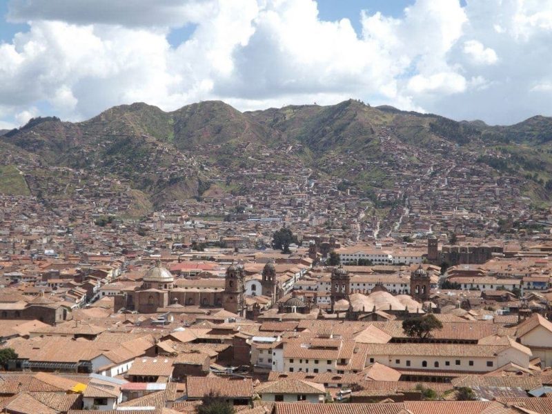 looking across the city of cusco towards large brown mountains on a cloudy day - the city centre is densely populated with buildings with mostly reddish terracotta tiled roofs. 