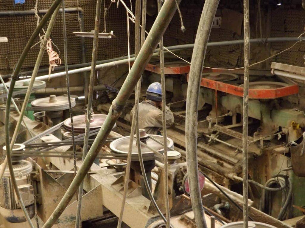 Miner in grey overalls and a blue helmet sitting amidst mining machinery and various pipes. 