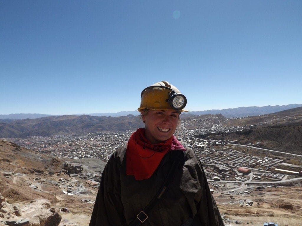 Emily wearing grey overalls, a red bandana and a yellow mining helmet with a large headtorch on top. She is smiling at the camera with an aerial view over Potosi city skyline behind. 