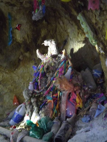 Blurry photo of a grey rocky sculpture of El Tio, the god of the mine, surrounded by colouful paper bunting and gifts of coca leaves, alcohol and cigarettes