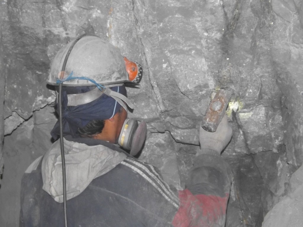 Close up of a miner hitting a rock wall with a hammer. He is wearing a black Adidas jacket and a grey helmet.