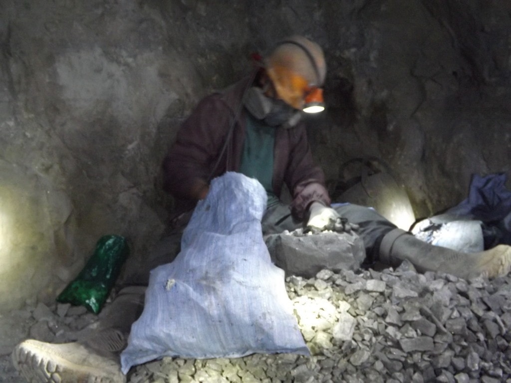 Miner at work inside the Potosi mine, he is wearing grey trousers and a dark red jacket with a yellow helmet. He is sitting down next to a rock wall sorting stones into a blue bag. 