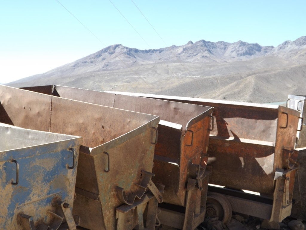 Rusted metal mine carts in a row with a view of brown rocky mountains behind and clear blue sky above. 