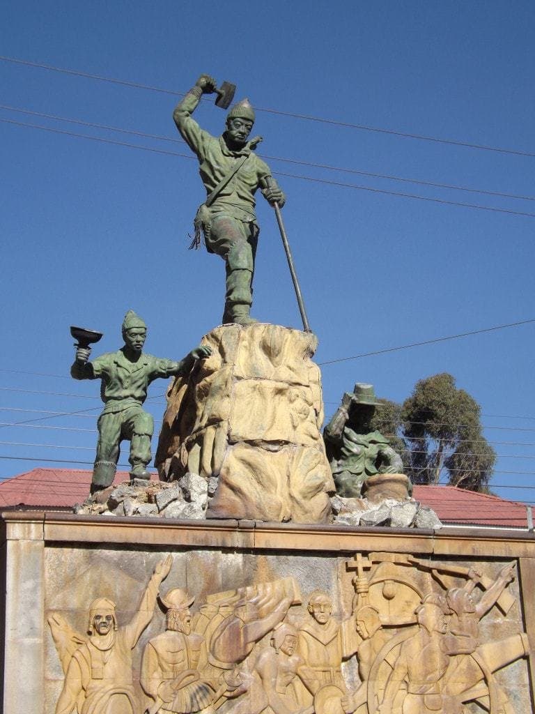 Bronze statue of three miners using hammers and picks, standing on a stone plinth with carvings of miners, seen against a clear blue sky