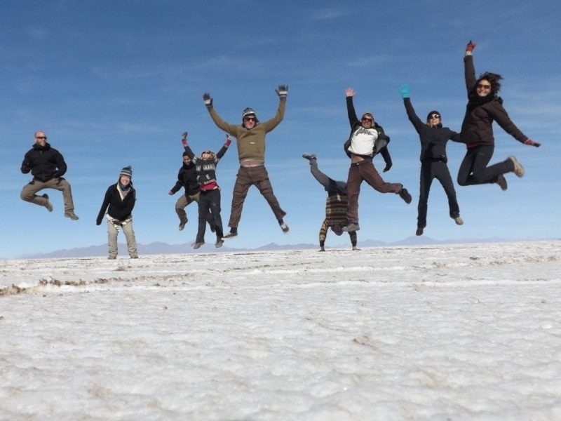 group of people in winter outdoor clothing jumping into the air with arms up above the white salt desert at salar de uyuni in bolivia against a blue sky