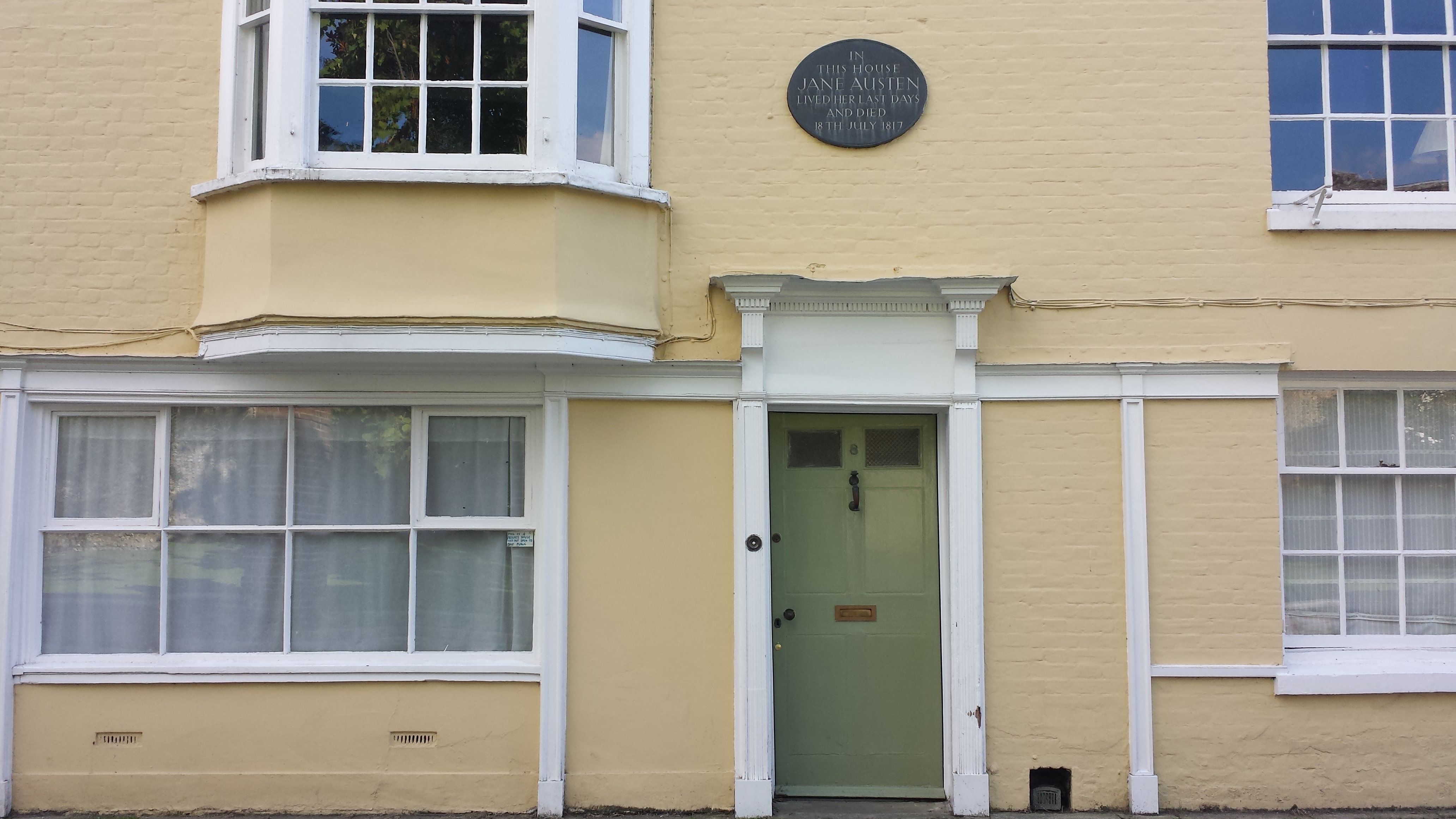 Facade of a small stone cottage with yellow painted walls and a fgreen front door, above the door a bronze plaque reads "in this house hane austen lived her last days and died"