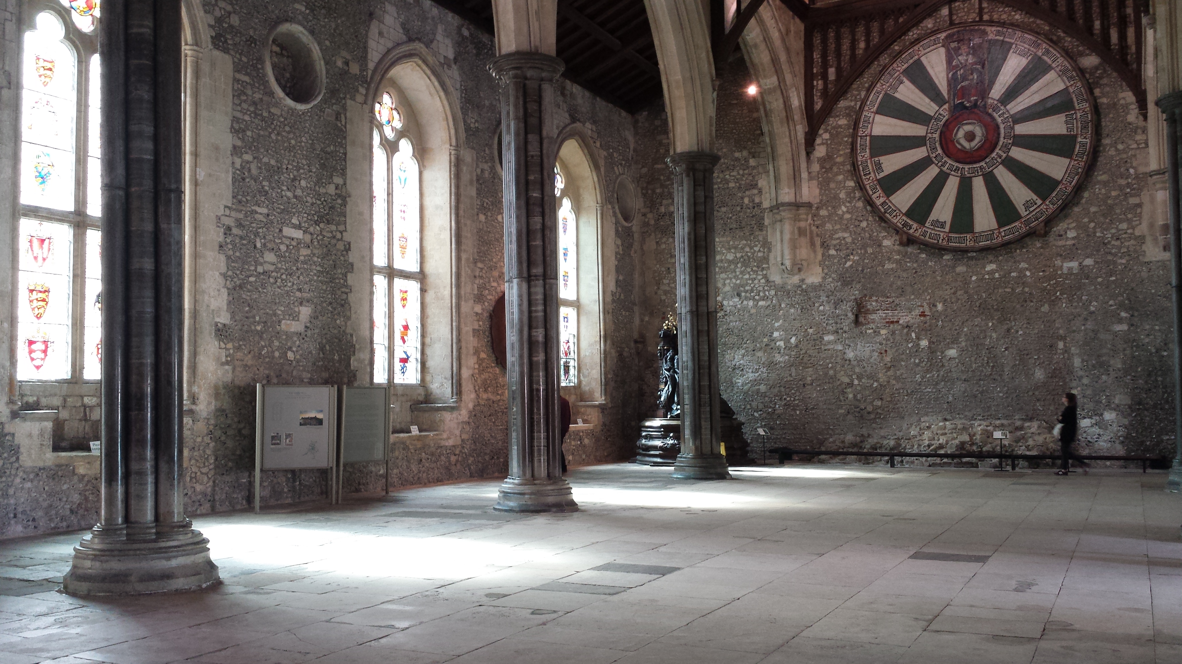 Interior of The Great Hall in Winchester with stone pillars and grey flagstone floors and stone walls. There is a green and white painted Round Table hung on the wall. 