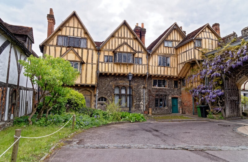 exterior of a historic building with stone walls on the first floor and yellow painted timber clad walls on the top half in a courtyard with purple wisteria growing over the walls