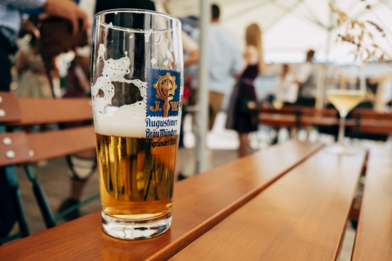 close up of a half full pint glass of beer on a woden table with the interior of a traditonal German beer tent out of focus in the background. Where to celebrate Oktoberfest in the UK. 