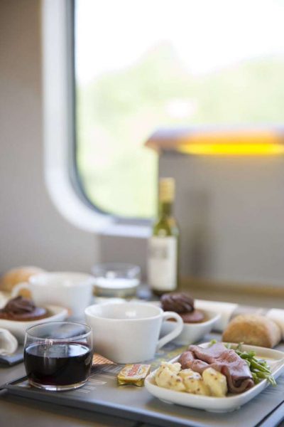 A meal on a grey tray with a small tumbler of wine, a white coffe cup, and a white square plate with potatoes ham and green beans, all on a table on a train with the train window in the background. Eurostar Standard vs Standard Premier - onboard meal