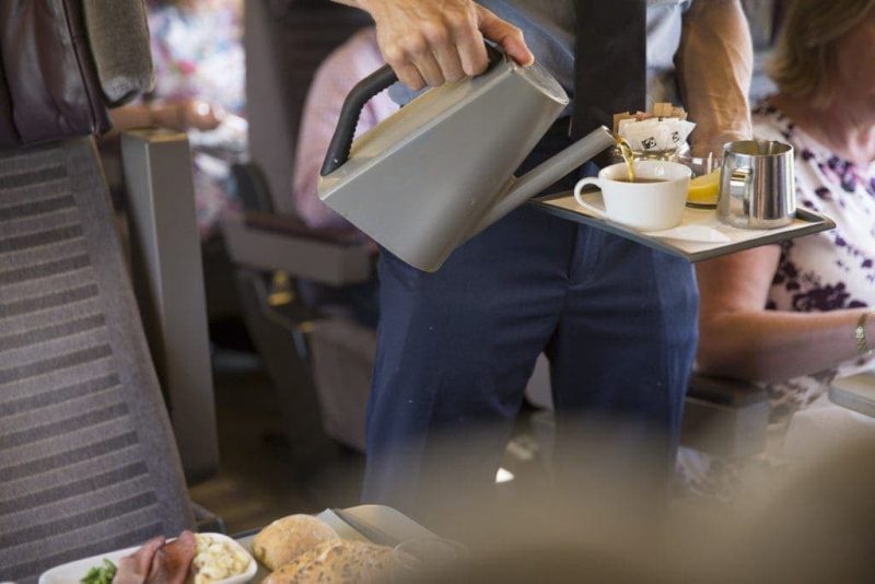 Lower half of a waiter wearing blue trousers pouring coffee from a kettle into a white mug on a tray on board a train. Eurostar Standard vs Standard Premier - interior of premier coach