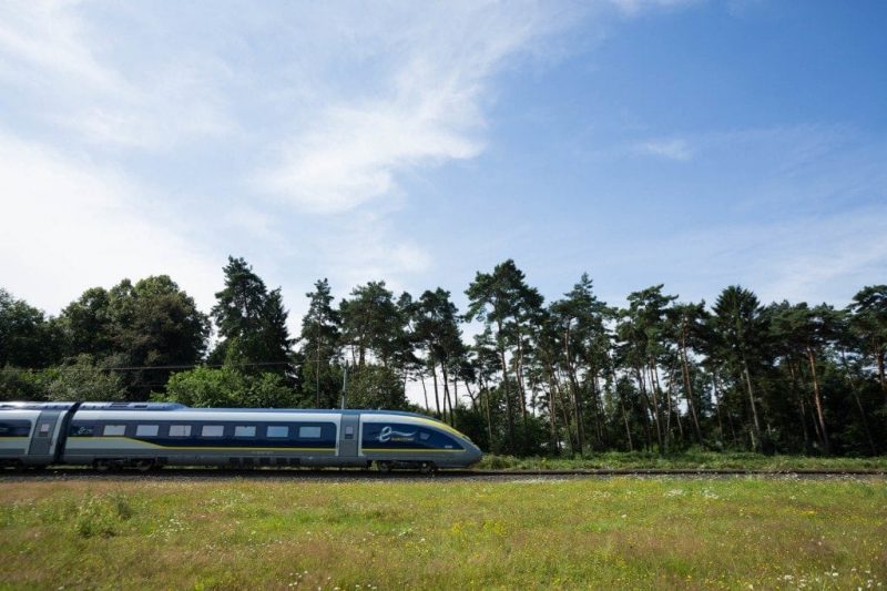 Eurostar train running along a track with a grassy field in front and a row of tall green trees behind