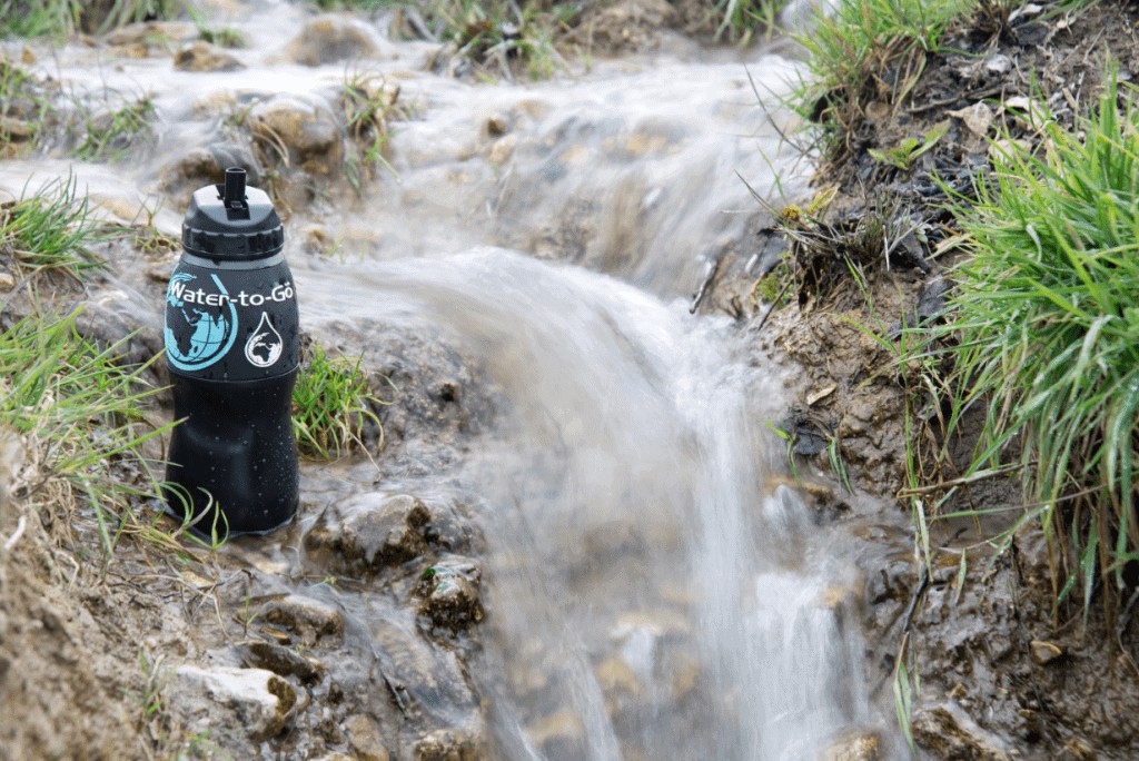 Black bottle with the name Water-to-Go printed on the side next to a small stream surrounded by grass.
