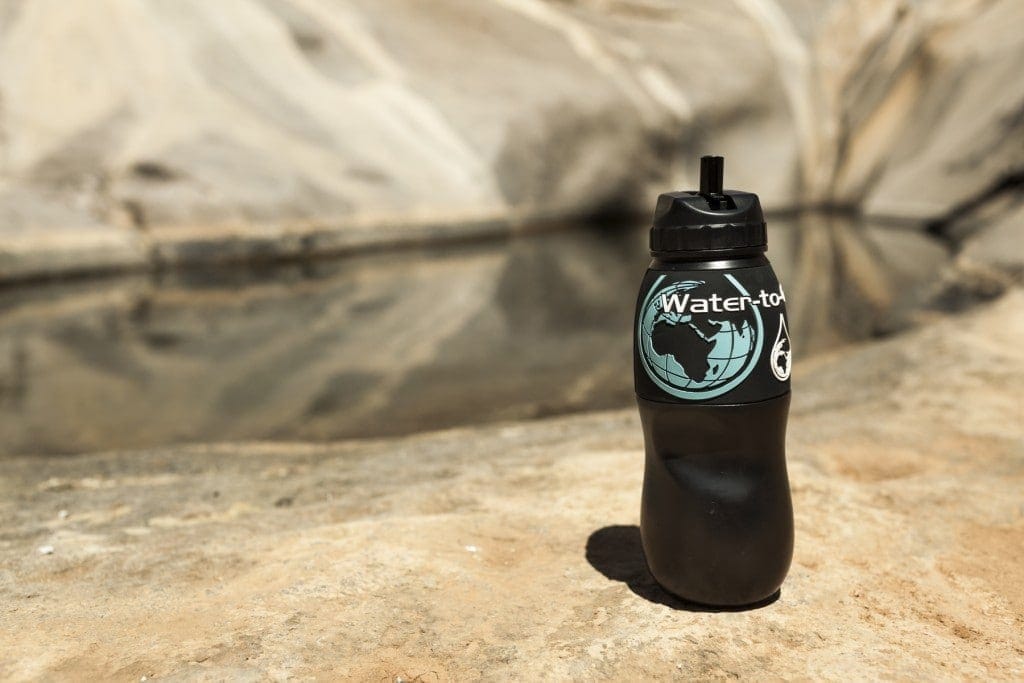 Black water bottle with the name Water-to-Go printed on the sleeve above a globe logo, on a rocky ledge with a pond out of focus behind.