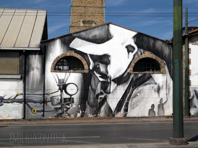 Black and white street art mural on the Athens gas factory with part of the chimney visible behind