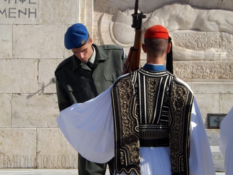 Changing of the Guard at the Tomb of the Unknown Soldier in Athens, the guard is wearing a white tunic with long sleaves and a patterned shawl over the top, with a small red hat on his head. He has his back to the camera and an officer in uniform is inspecting his sleave. 