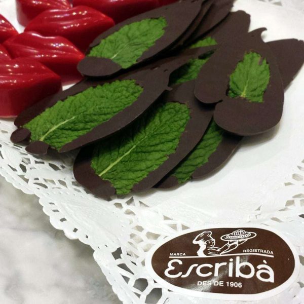 Close up of a white paper napkin with chocolate coated mint leaves on it and red lip-shaped chocolates with a brown sticker on the corner of the napkin with the shop name Escriba on it. Chocolate tour barcelona