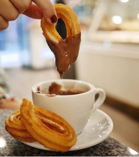 Close up of a white mug filled with melted cholcate on a saucer with two curled churros. A hand with red nail polish is lifting a chocolate covered churro out of the cup. Best chocolate in Barcelona.