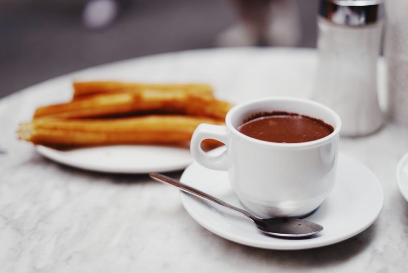 white marble table top with a white mug filled with melted chocolate on a white saucer next to a side plate with several long sticks of churros on it