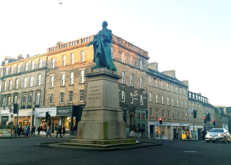 Blue coloured copper statue of George IV on a beige stone plinth in the middle of a crossroad with 4-storey beige stone terraced townhouses behind and high street shops along the ground floor. Unusual Things to do in Edinburgh New Town