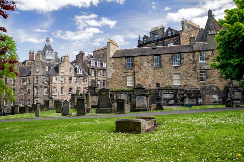 church graveyard with a grassy lawn covered in daisies with many grey stone gravestones. behind the graveyard there are beige-coloured stone terraced houses with grey slate roofs. 