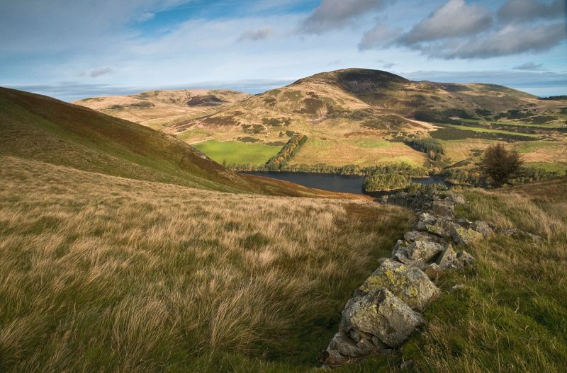 Landscape in the Pentland Hills in Scotland wiht a grassy slope leading down to a dark grey lake at the foot of a brownish grassy hill