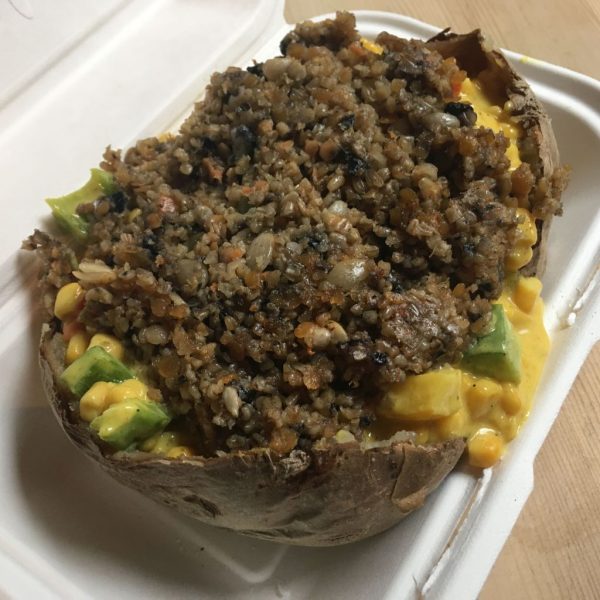 Close up of a jacket potato cut in half and topped with avocado, sweetcorn, a yellow sauce, and a thick stew of lentils and quinoa, inside a white polystyrene takeaway box from The Baked Potato Shop 