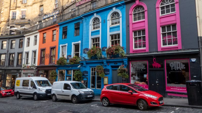 A cobbled street with a row of 3-storey terraced townhouses all painted very birght colours (white, orange, blue, light blue, and grey with hot pink trim). Unusual Things to do in Edinburgh