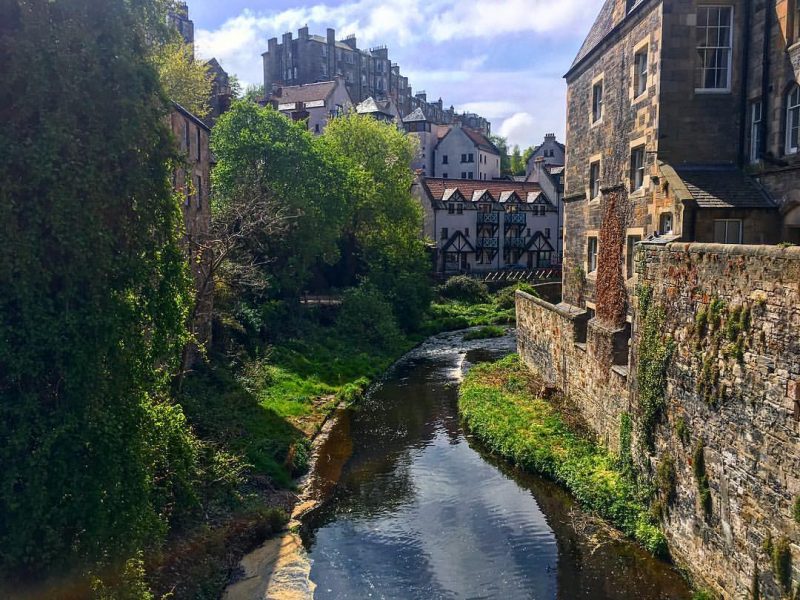 Looking from a bridge down at a river called the Water of Leith in Edinburgh, with tall stone houses on either side and more old houses on a hilltop in the distance.