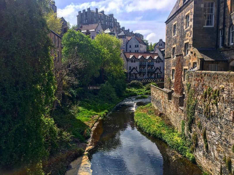 Looking from a bridge down at a river called the Water of Leith in Edinburgh, with tall stone houses on either side and more old houses on a hilltop in the distance. 