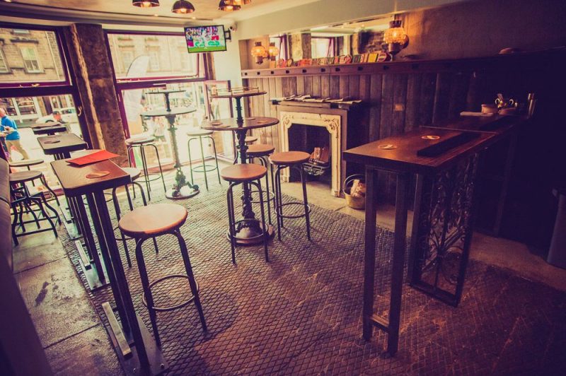 Interior of the Wee Pub, the Smallest pub in Scotland, wiht wooden bar stools around high wooden tables. the floor is stone tiles and the wall is covered with dark wood panelling with a stone fieplace.